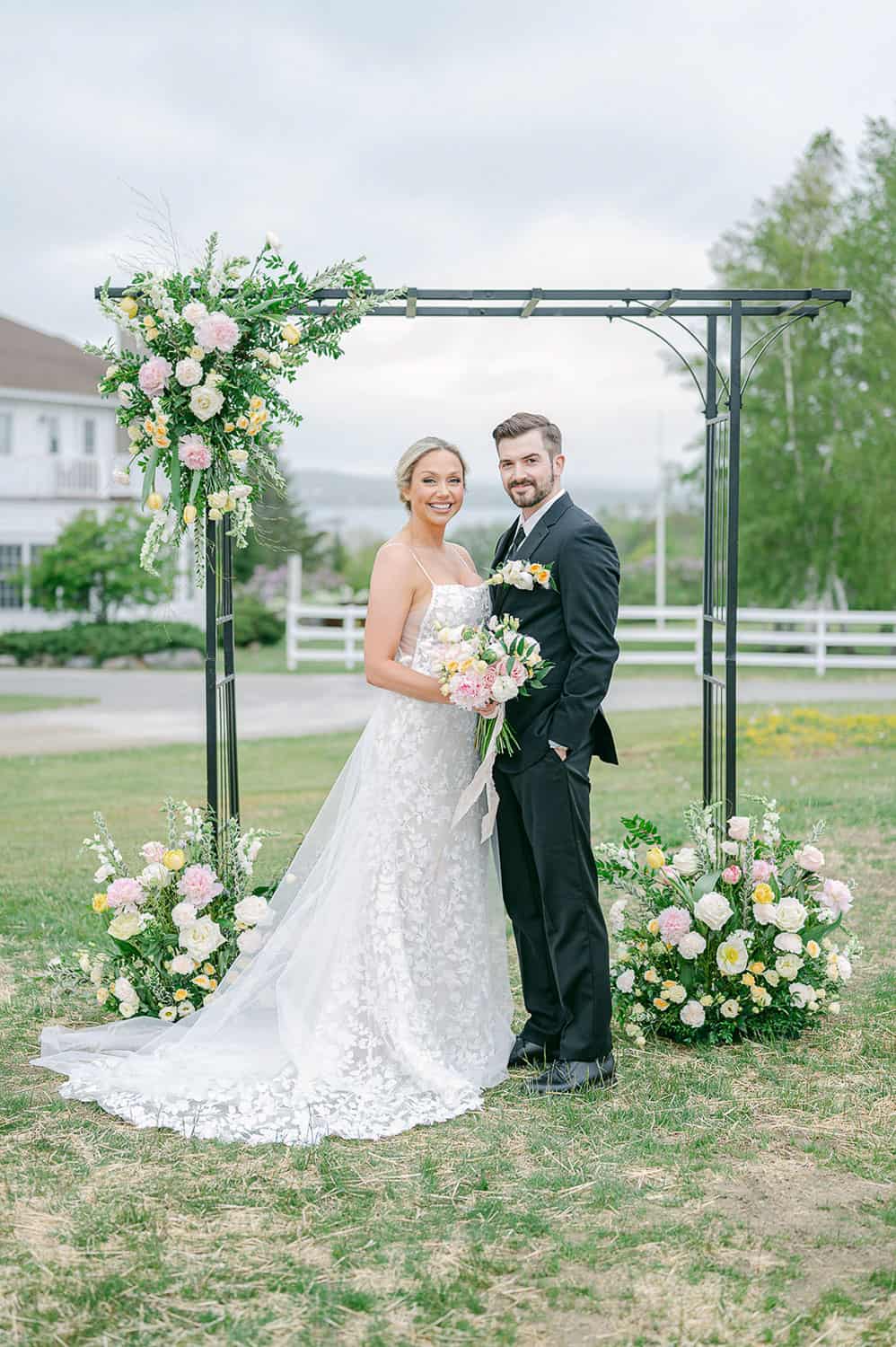 A bride with a bouquet and a groom smiling under a floral arch in a field, with a house in the background.