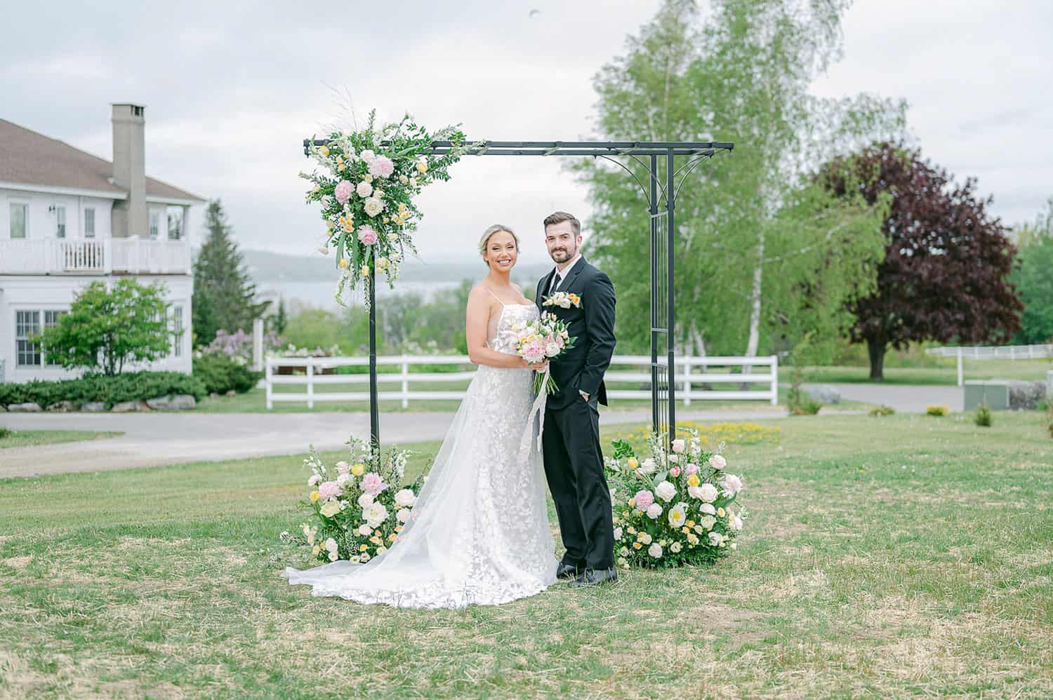 A bride with a bouquet and a groom smiling under a floral arch in a field, with a house in the background.