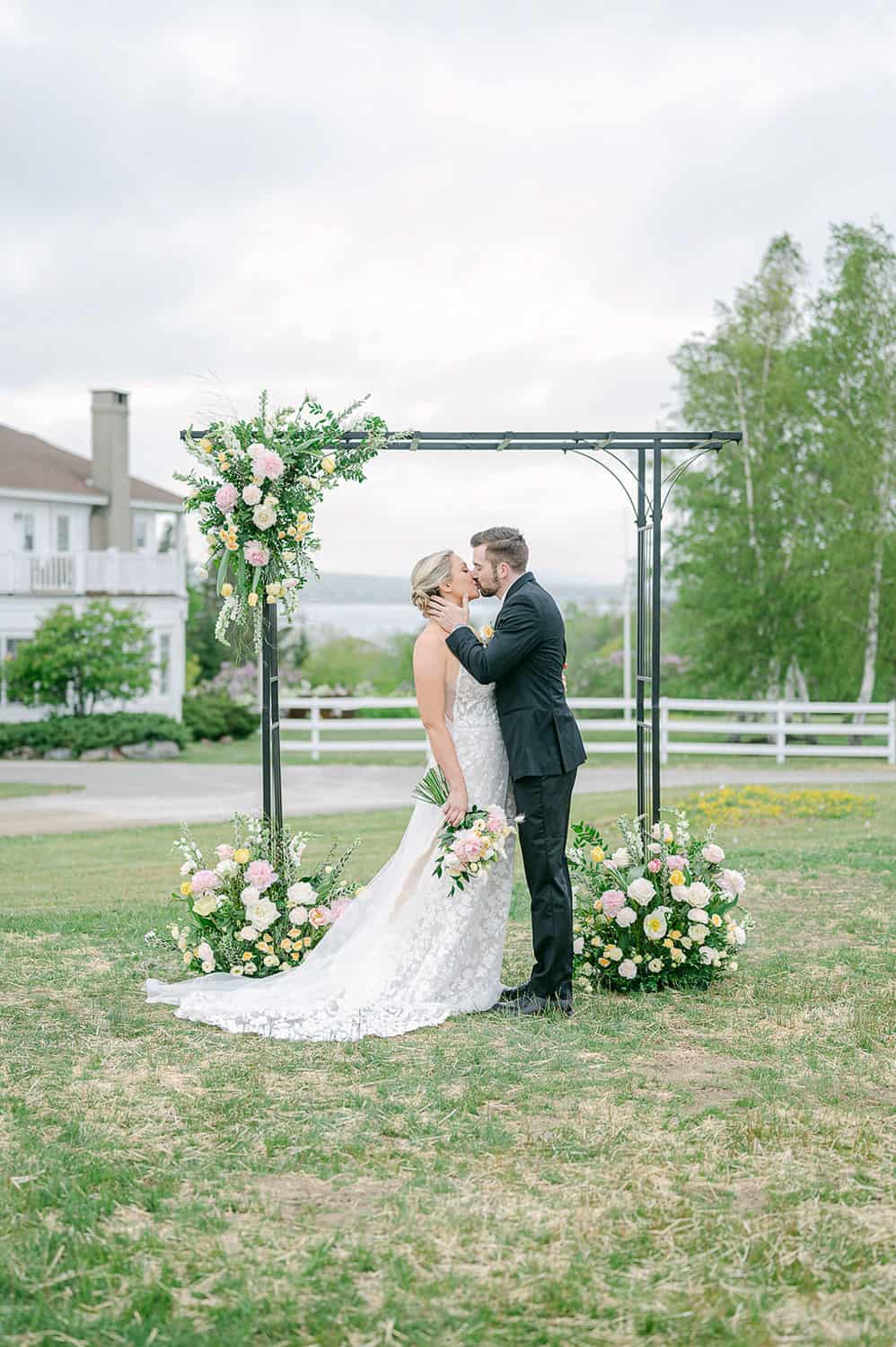 A bride with a bouquet and a groom kissing under a floral arch in a field, with a house in the background.