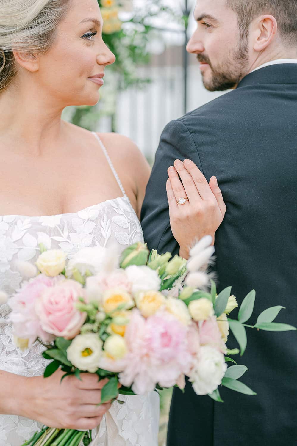 Bride holding a bouquet of pink and yellow flowers gazing at her groom in a garden setting.