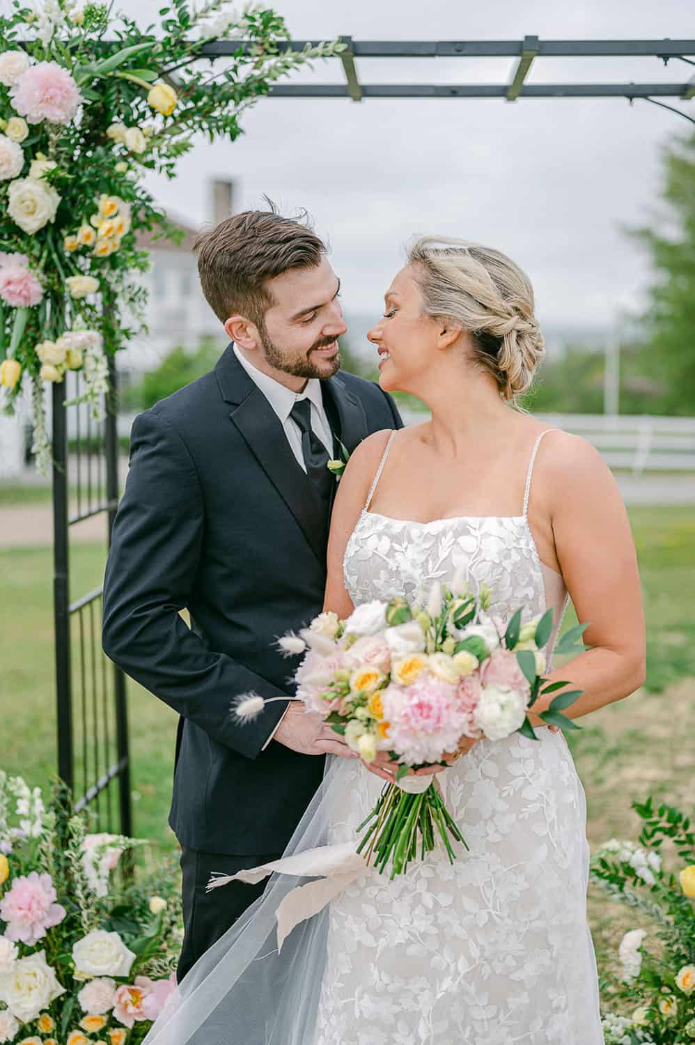 A bride and groom smiling at each other under a floral arch, with the bride holding a bouquet, in an outdoor setting.