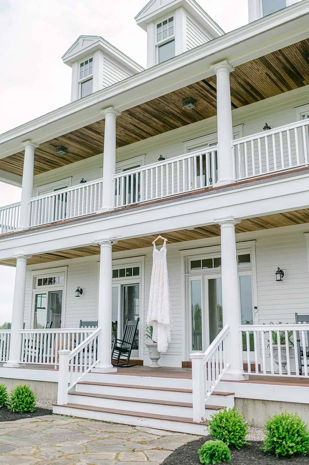 A white wedding dress hanging from the balcony of an elegant two-story house with a covered porch and white columns.