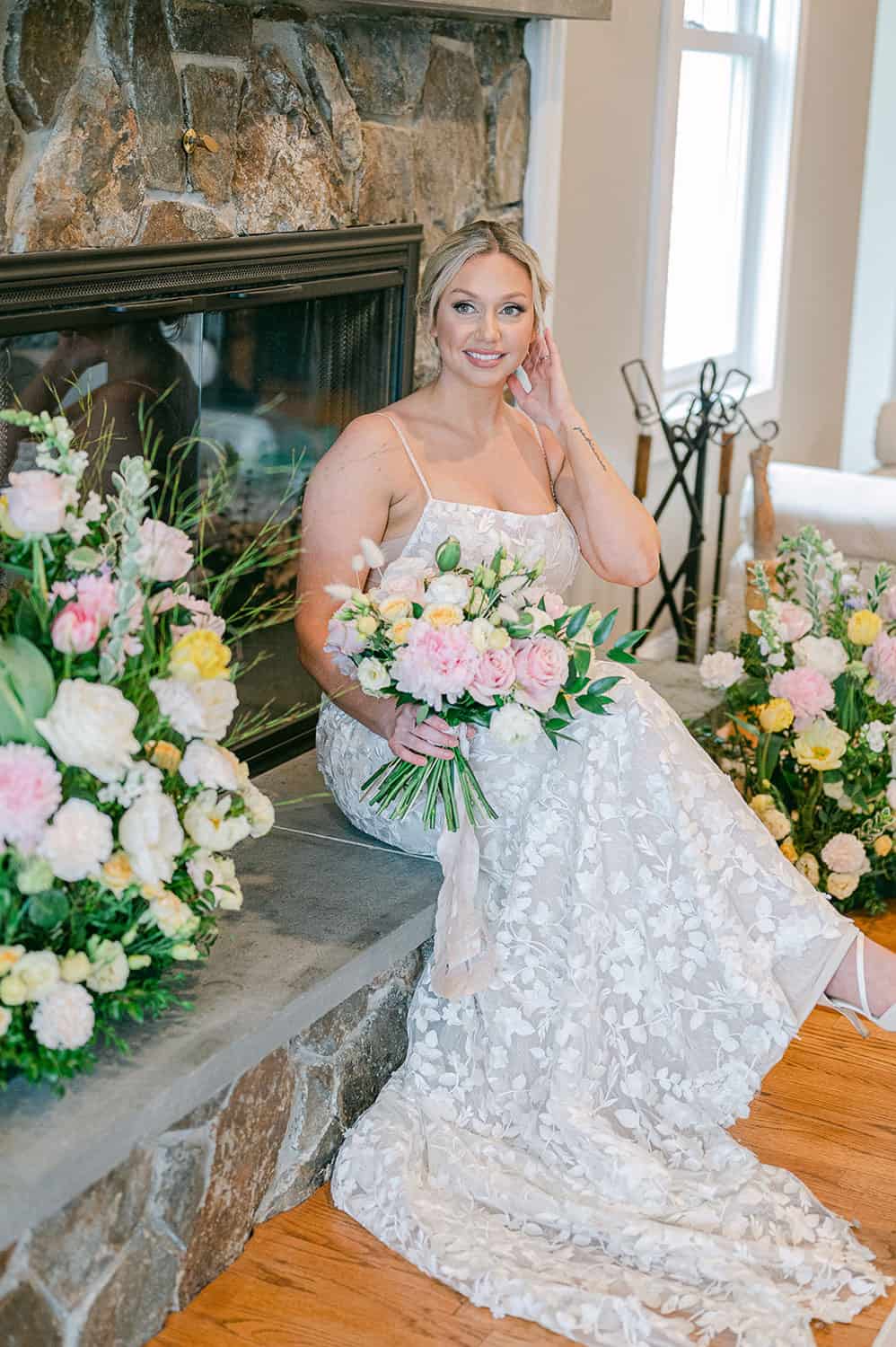 A bride in a lace wedding gown sitting by a fireplace, holding a bouquet of pink and white flowers, smiling at the camera.