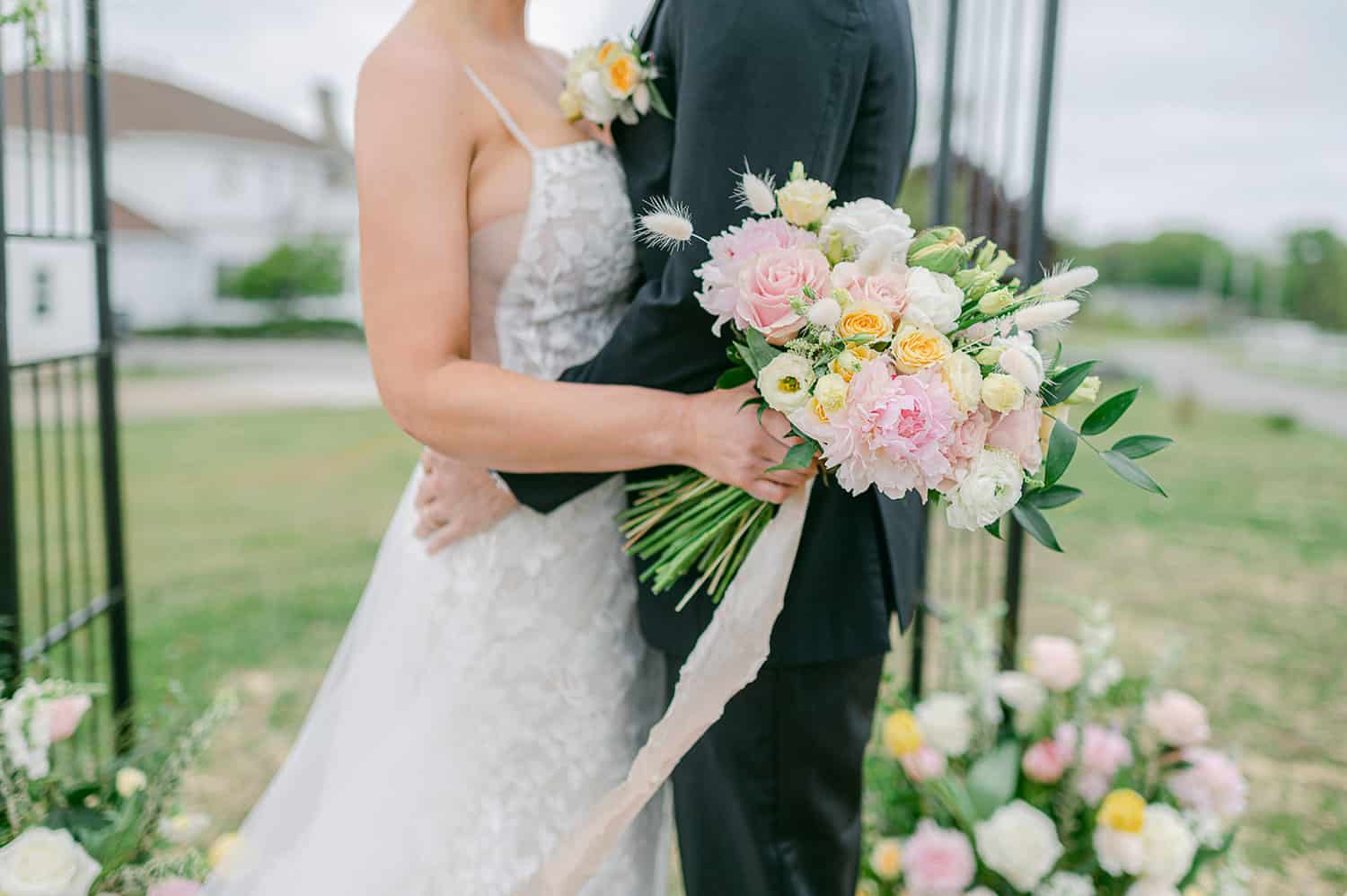 Bride and groom holding a bouquet of pastel flowers, close-up with a soft focus background in a garden setting.