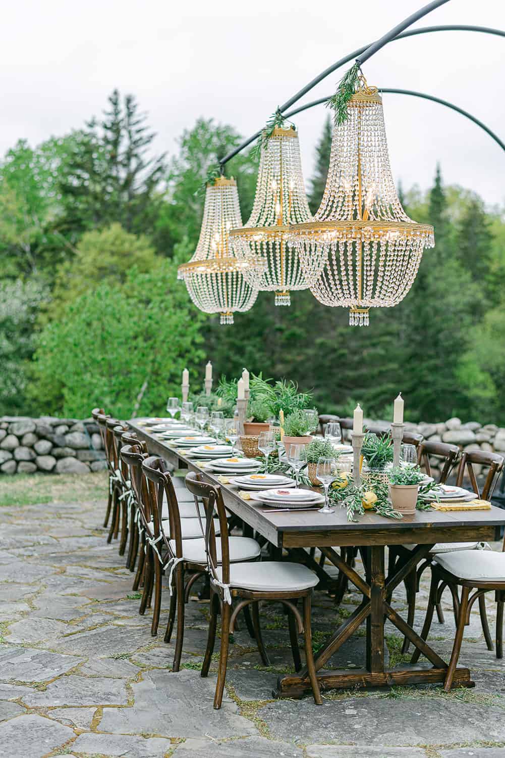 An elegantly set outdoor reception table with beaded chandeliers hanging above, amidst a lush garden setting.