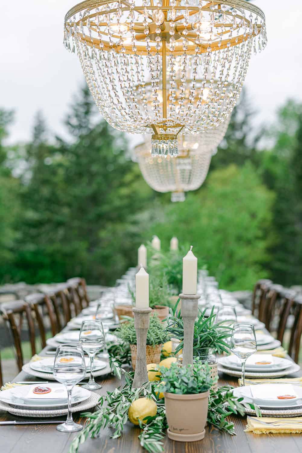 A close up of an elegantly set outdoor reception table with beaded chandeliers hanging above, amidst a lush garden setting.