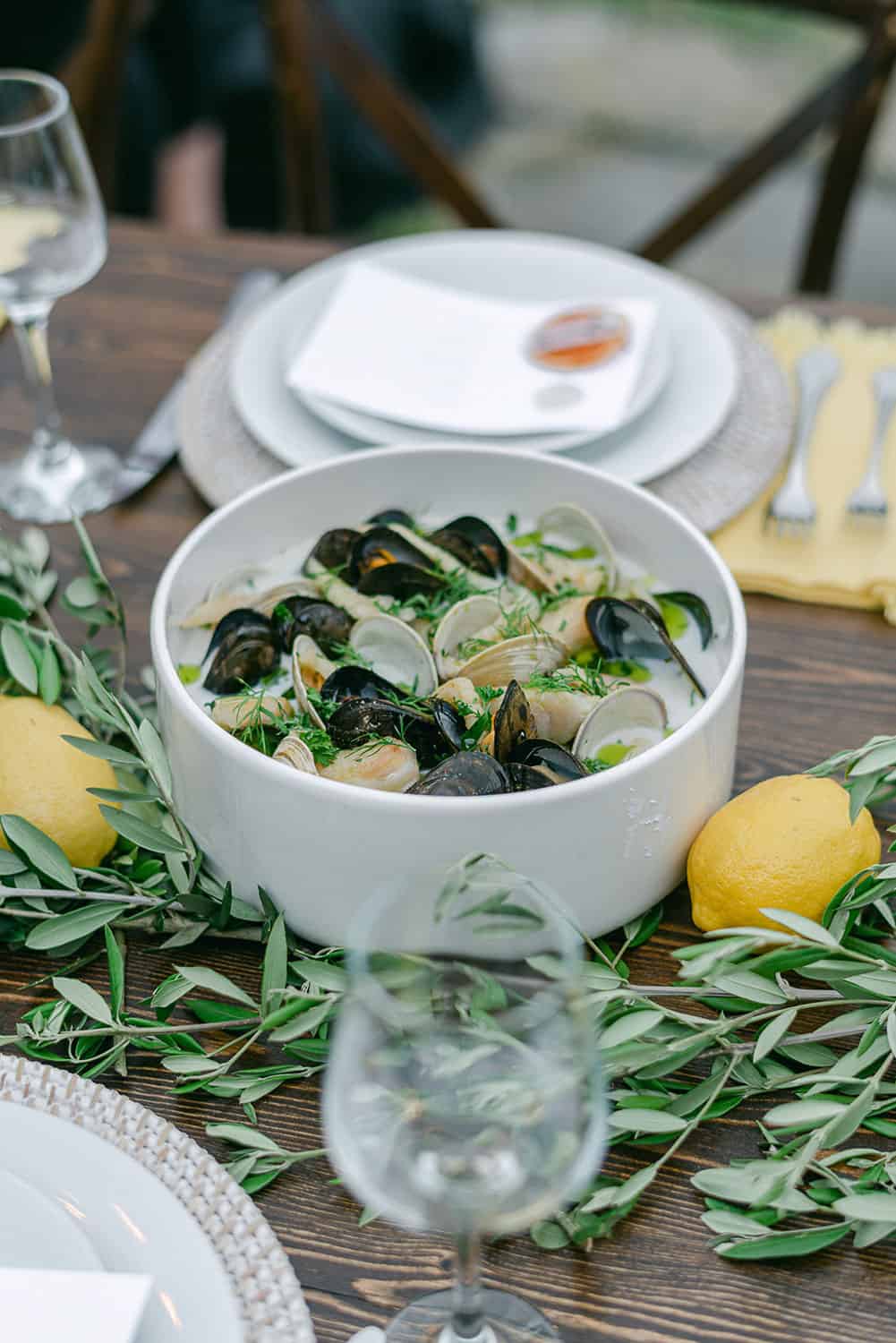 A bowl of mussels garnished with lemon and herbs on a reception table, accompanied by white plates, glassware, and silverware.