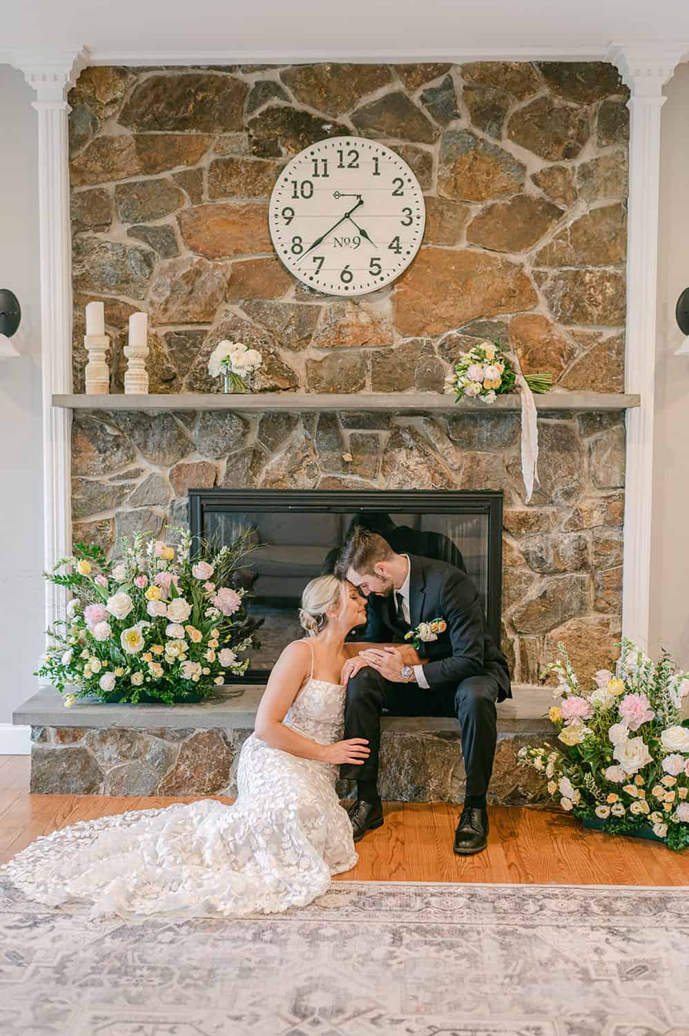 Bride and groom sitting by a stone fireplace, adorned with flowers and candles, sharing a moment under a large clock.