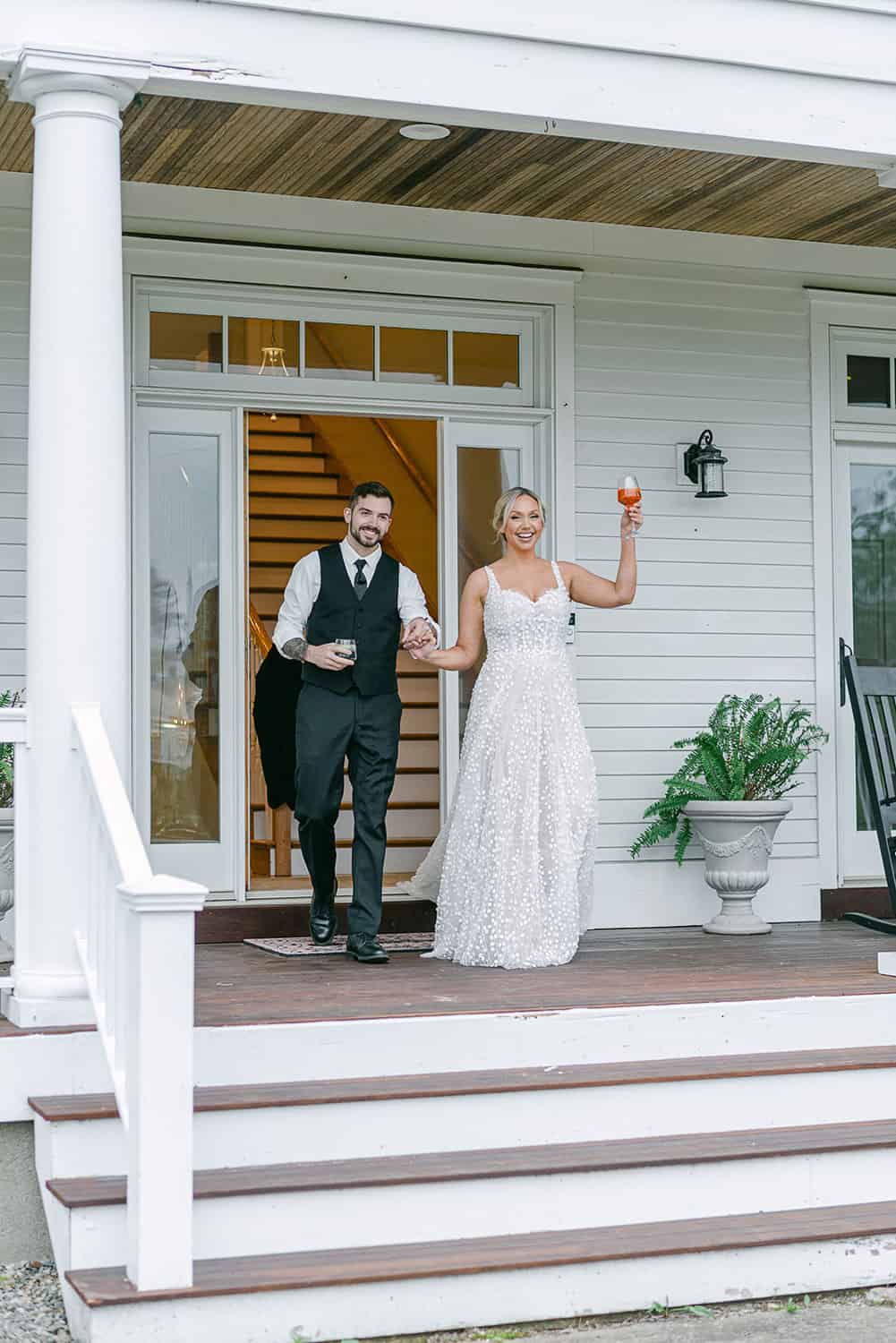 A bride and groom holding drinks, smiling and stepping out from a house onto the front porch.