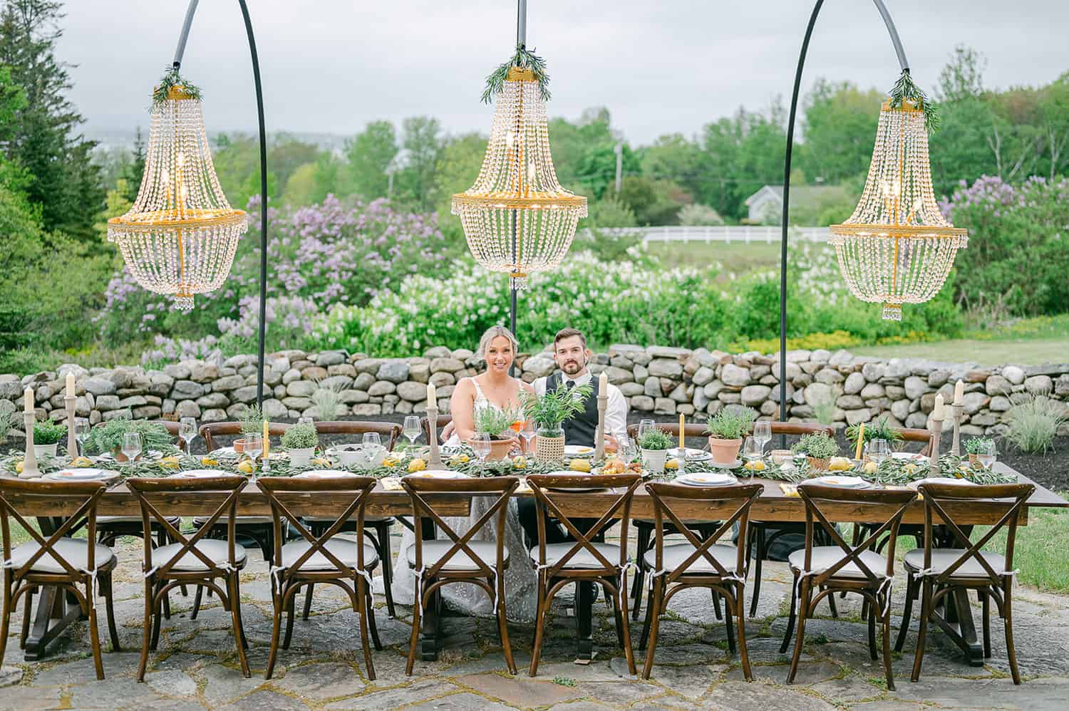 Bride and groom at an outdoor dining table with floral arrangements, under elegant chandelier lights, with a green backdrop.