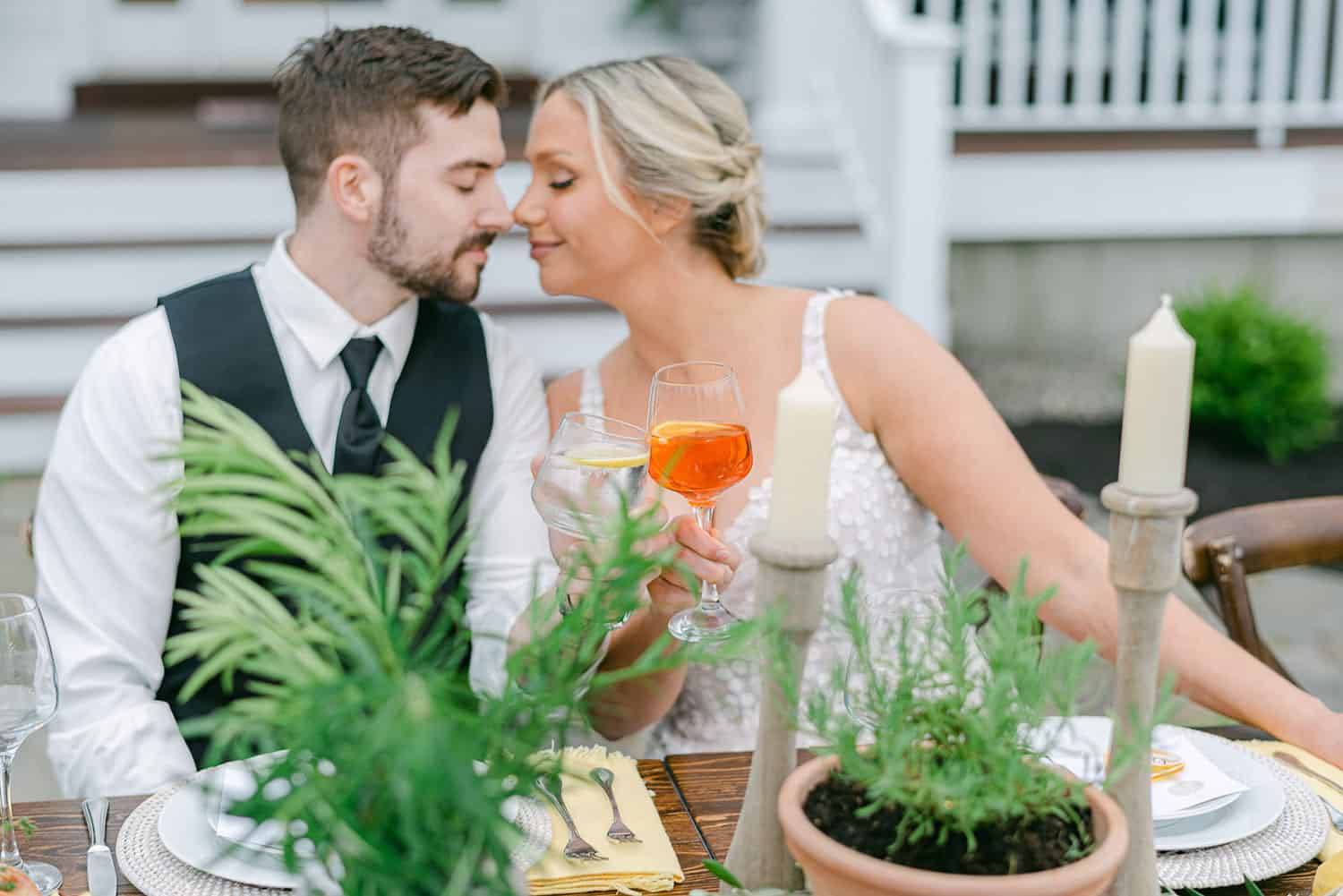 A couple toasting with drinks at an elegantly set outdoor dining table, leaning in for a kiss.