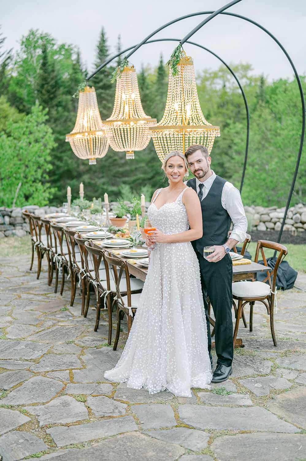 A bride and groom stand smiling together at an elegantly set outdoor wedding reception table under ornate chandeliers.