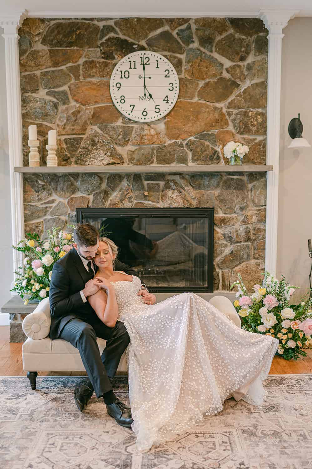 A bride and groom sitting closely on a white sofa in front of a stone fireplace, with the bride's gown spread out elegantly.