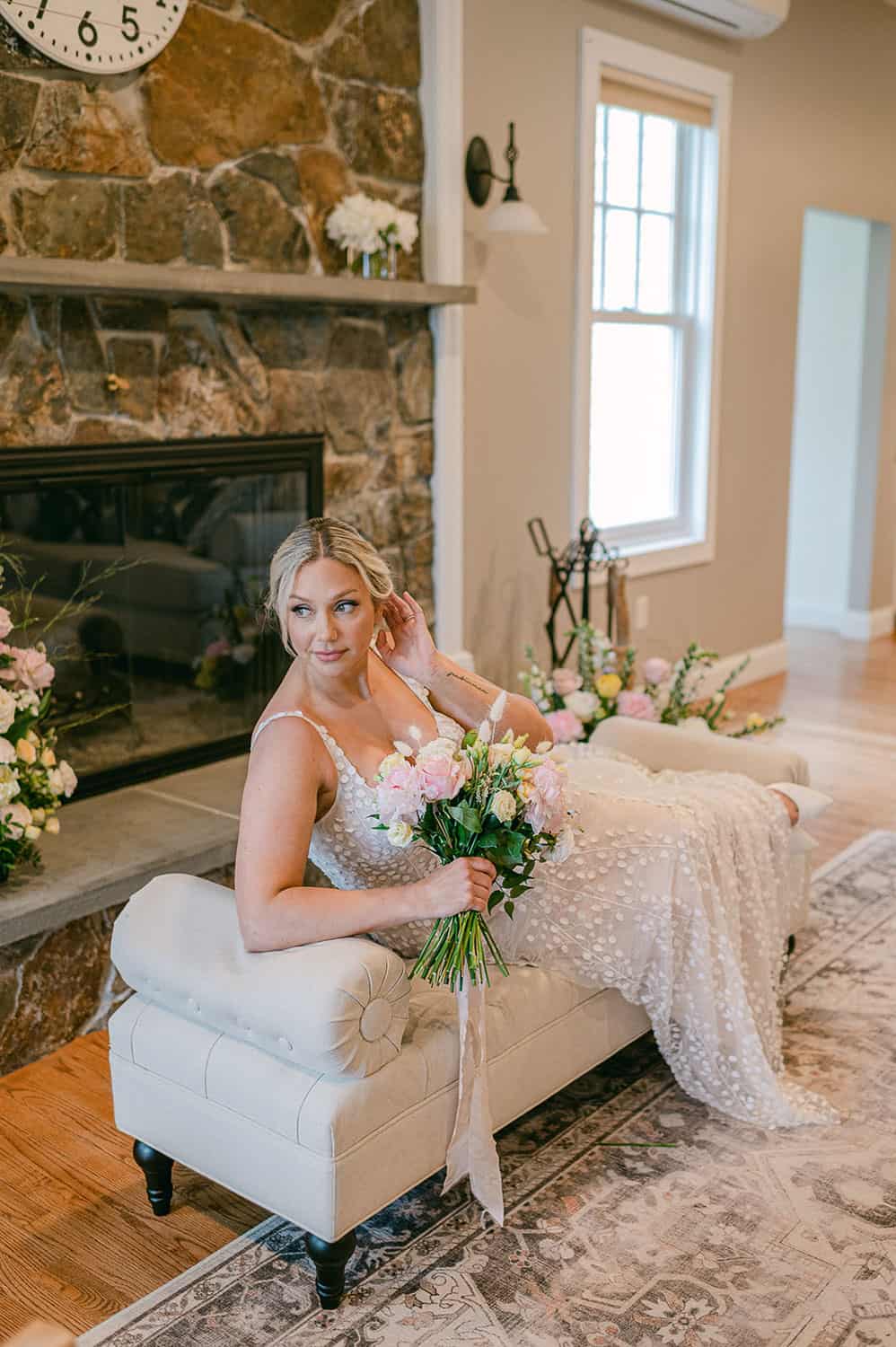 A bride in a white dress on a beige ottoman, holding a bouquet, with a fireplace and floral decorations in the background.