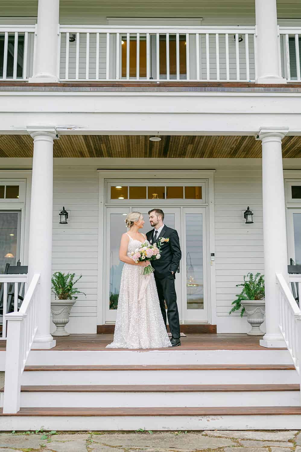 A bride and groom sharing a tender moment on the steps of a white house with a classic double-decker porch.