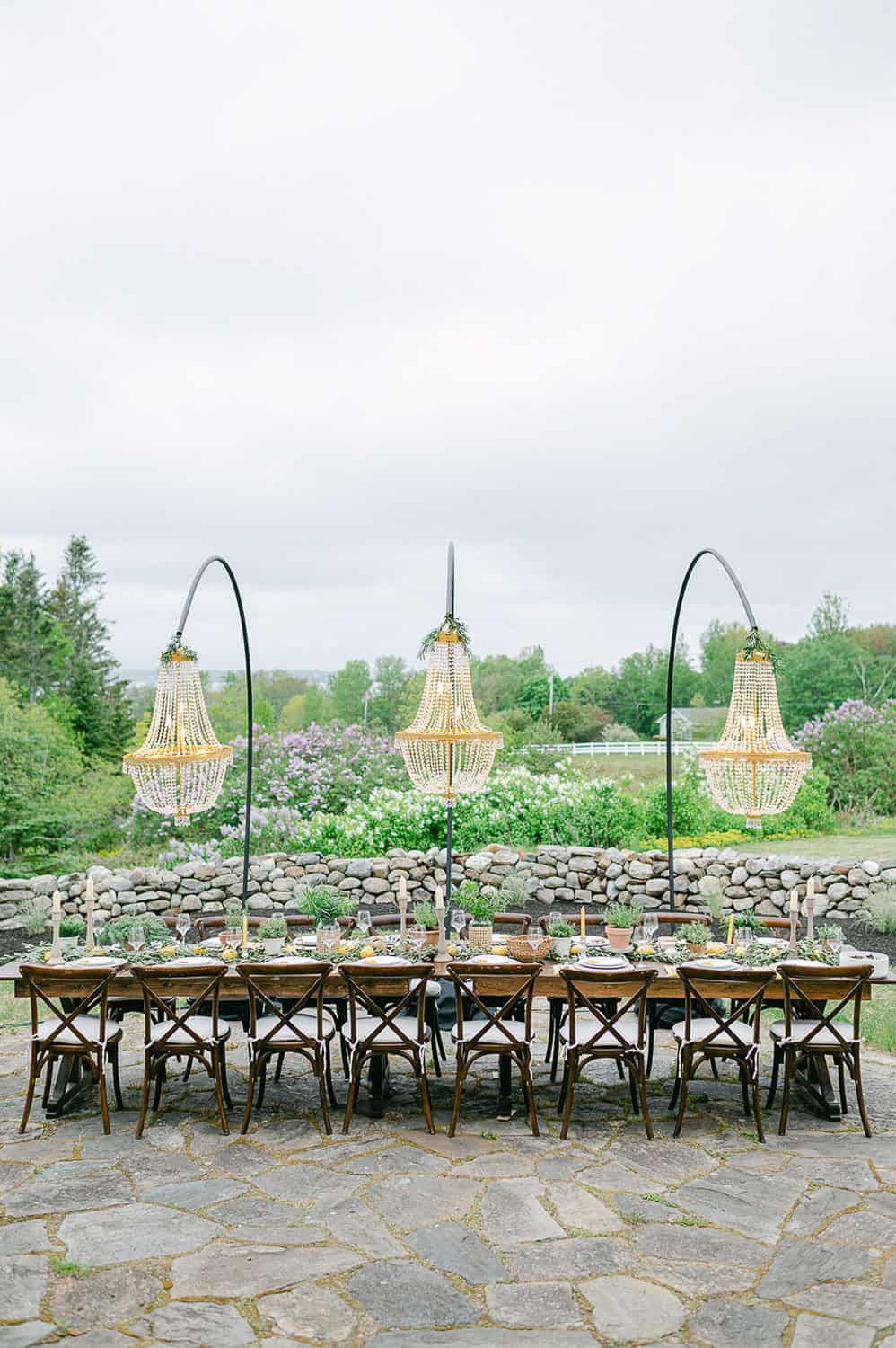 Outdoor table, with wooden chairs, and hanging basket lamps, on a stone patio surrounded by grass and a curved stone wall.