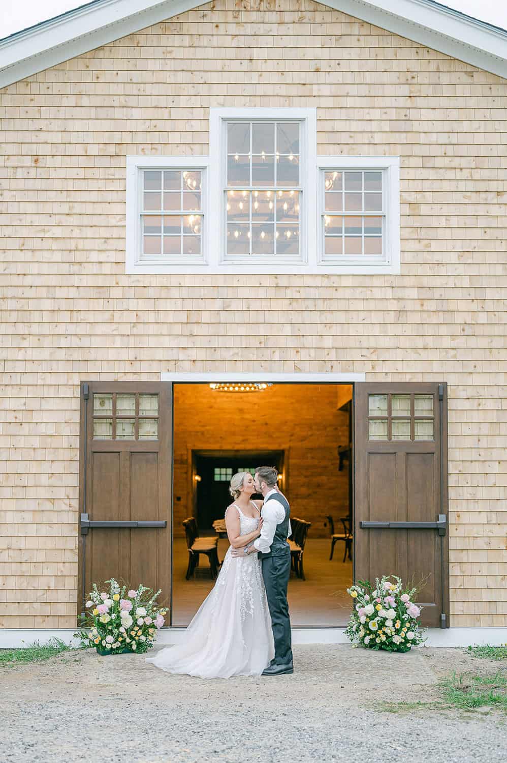 A couple embracing in front of an open barn door, adorned with flowers, under a string of lights inside the barn.