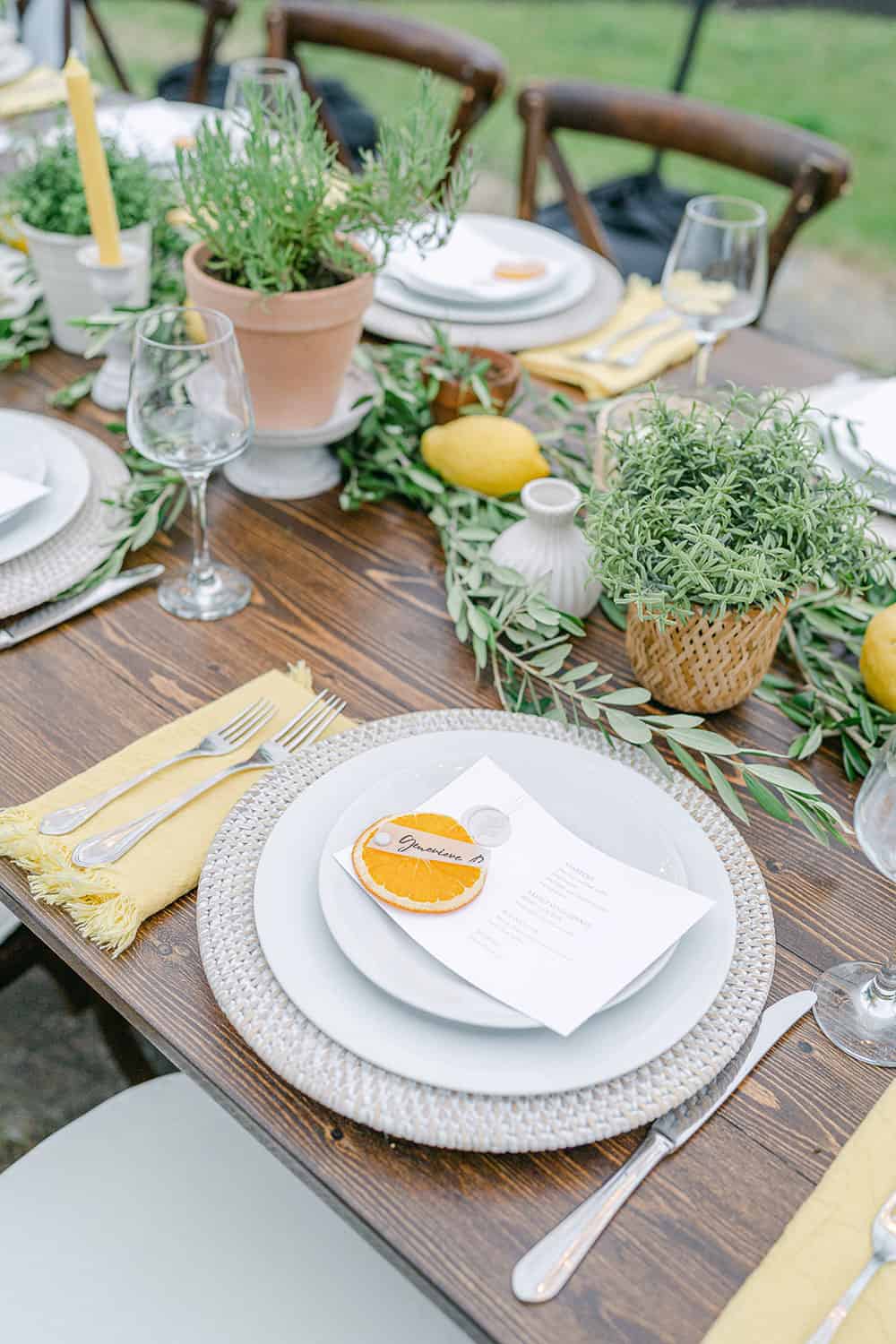 Reception table set with white plates, yellow napkins, and centered with potted green herbs and lemons.