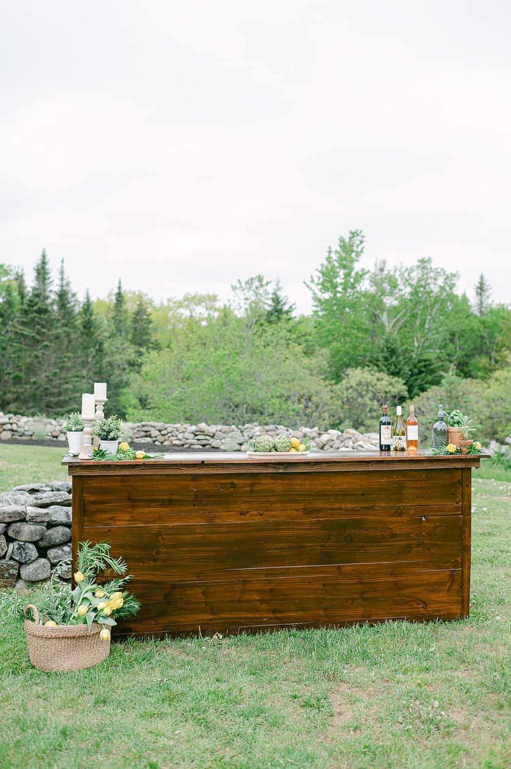 Outdoor wooden bar in a lush field with a pot of flowers beside it, surrounded by trees and a stone wall under a cloudy sky.