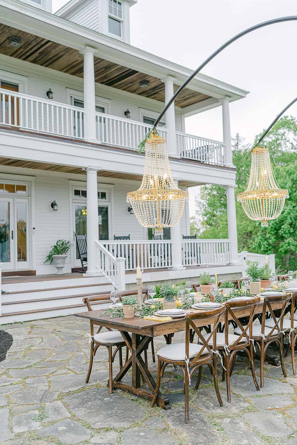 An elegant wooden reception table with rustic chairs, and hanging chandeliers, in front of a white house with a balcony.