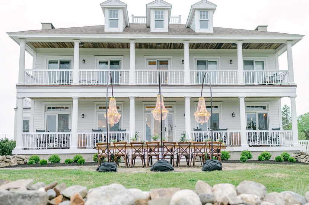 Private estate weddings in maine. An elegant wooden reception table with rustic chairs, and hanging chandeliers, in front of a white house with a two-story balcony.