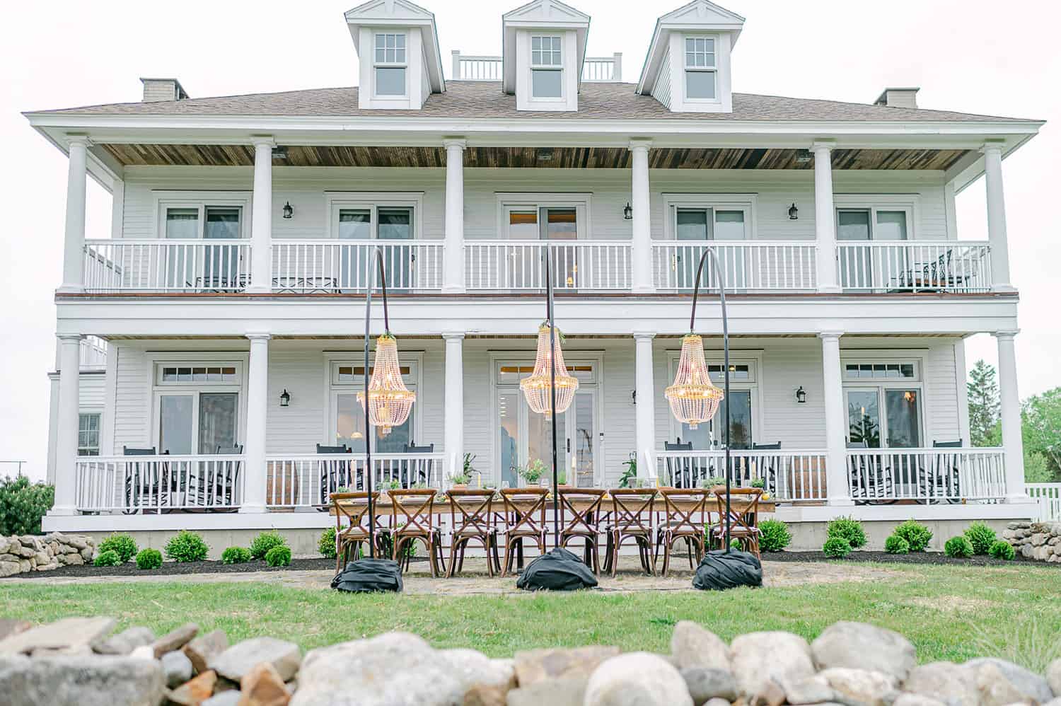 An elegant wooden reception table with rustic chairs, and hanging chandeliers, in front of a white house with a two-story balcony.