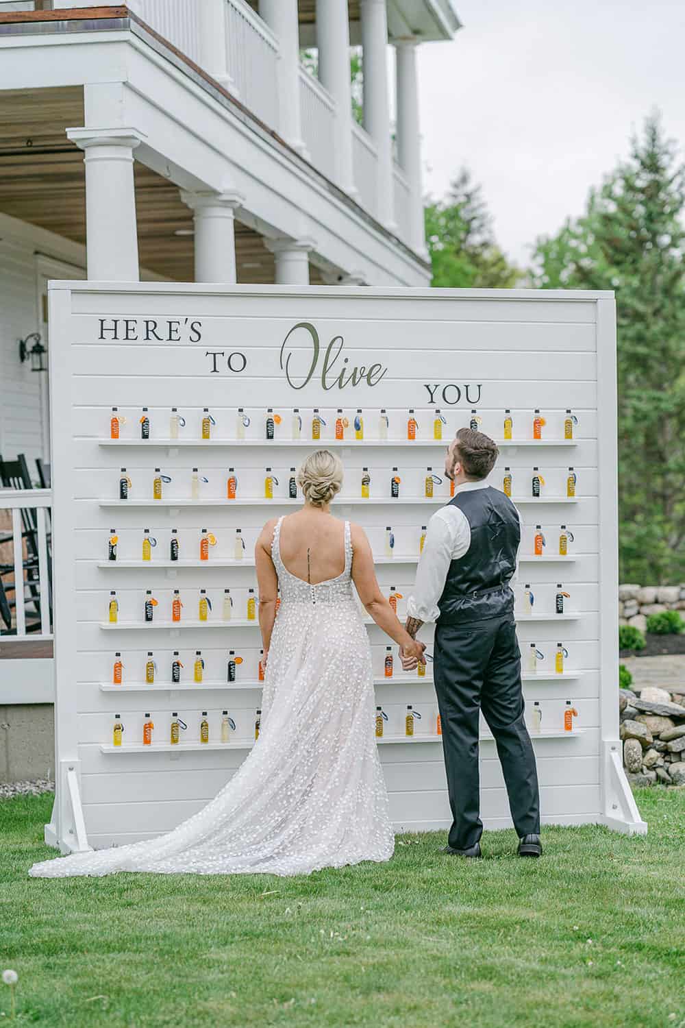 A bride and groom stand in front of a wall display of small labeled bottles, with "here's to olive you" featured at the top.