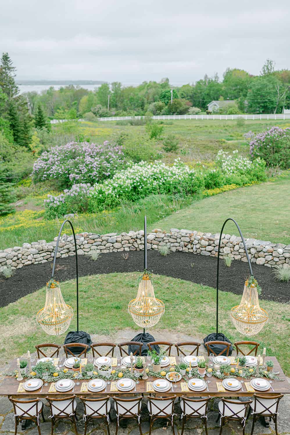 Outdoor table, with wooden chairs, and hanging basket lamps, on a stone patio surrounded by grass and a curved stone wall.