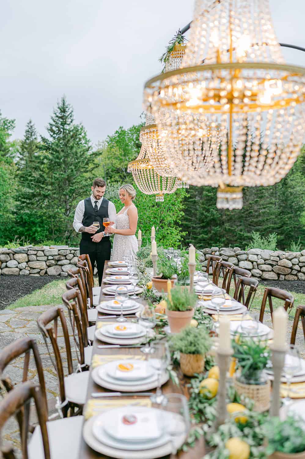 A bride and groom stand at the end of a beautifully set outdoor dining table under elegant chandeliers, surrounded by nature.