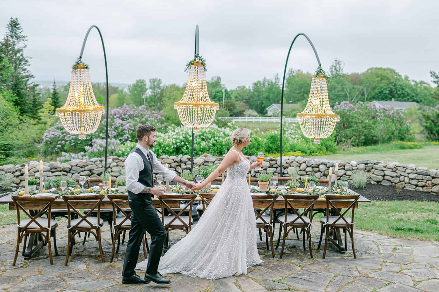 A bride and groom holding hands at an elegantly set long outdoor table with hanging chandeliers, surrounded by lush greenery.