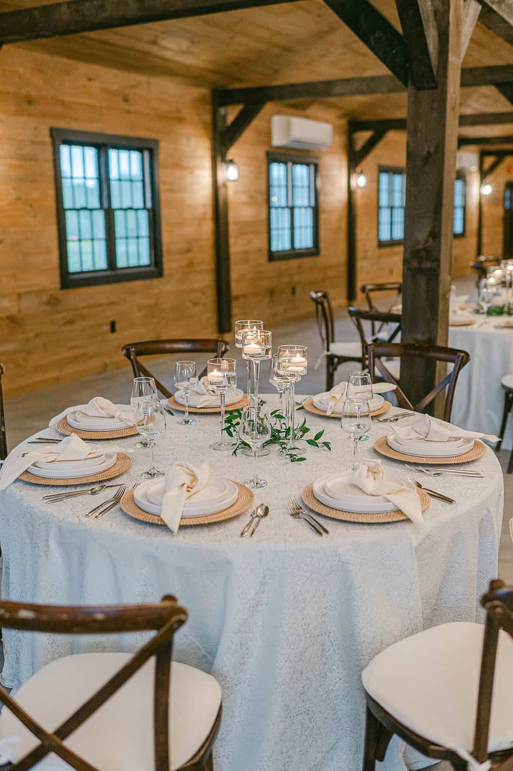Elegant wedding reception table decorated with plates, cutlery, and candles, surrounded by wooden chairs in a rustic venue.