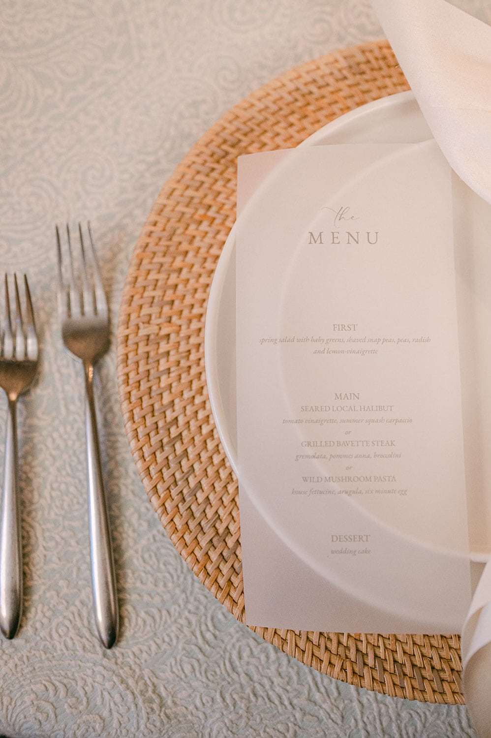 Table setting with a round wicker placemat, a white plate with a printed menu, and two forks, on a patterned tablecloth.