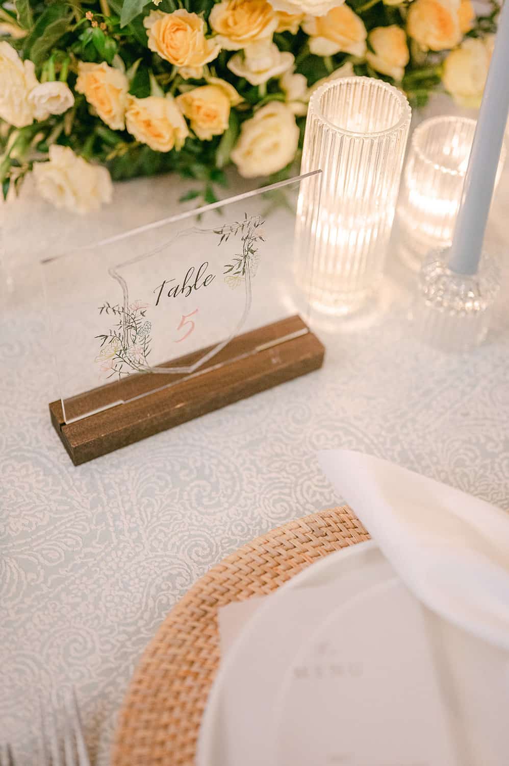 Elegant wedding table with a floral centerpiece, a "table 5" sign, candles, and fine dinnerware on a tablecloth.