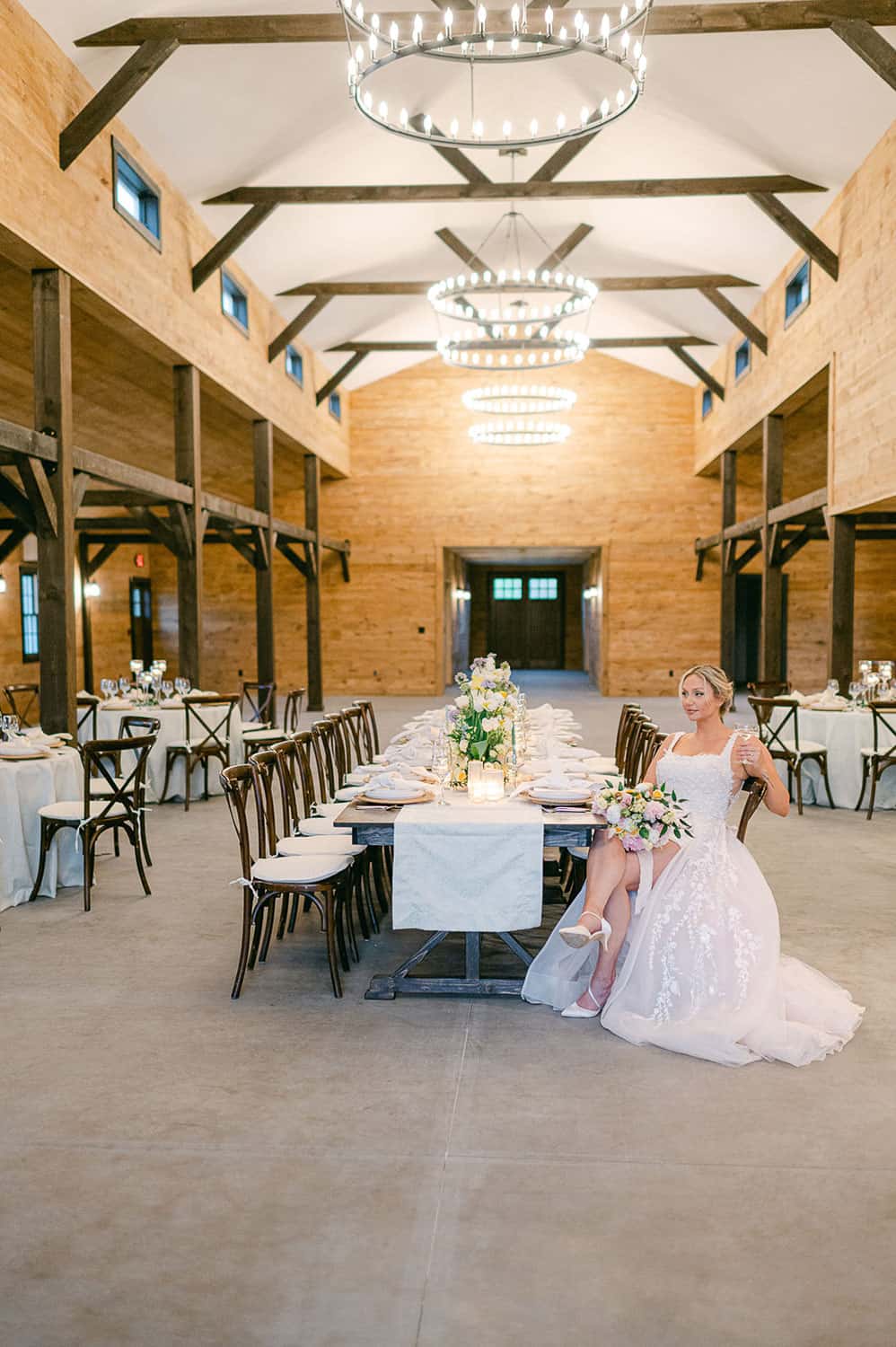 A bride in a white dress sits alone at a decorated banquet table in an elegant, rustic hall with wooden beams and chandeliers.