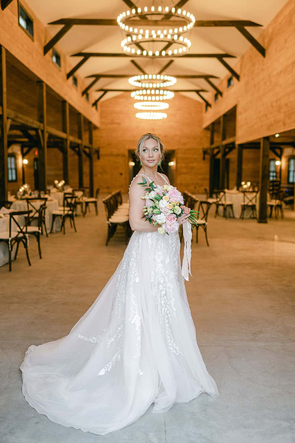 A bride in an elegant white gown holding a bouquet stands in a venue with wooden beams and chandeliers overhead.