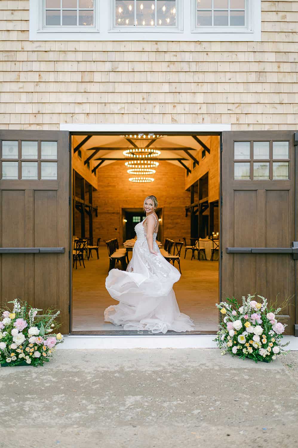 A bride twirls gracefully at the entrance of a barn with open doors, with floral arrangements, and a lit chandelier inside.