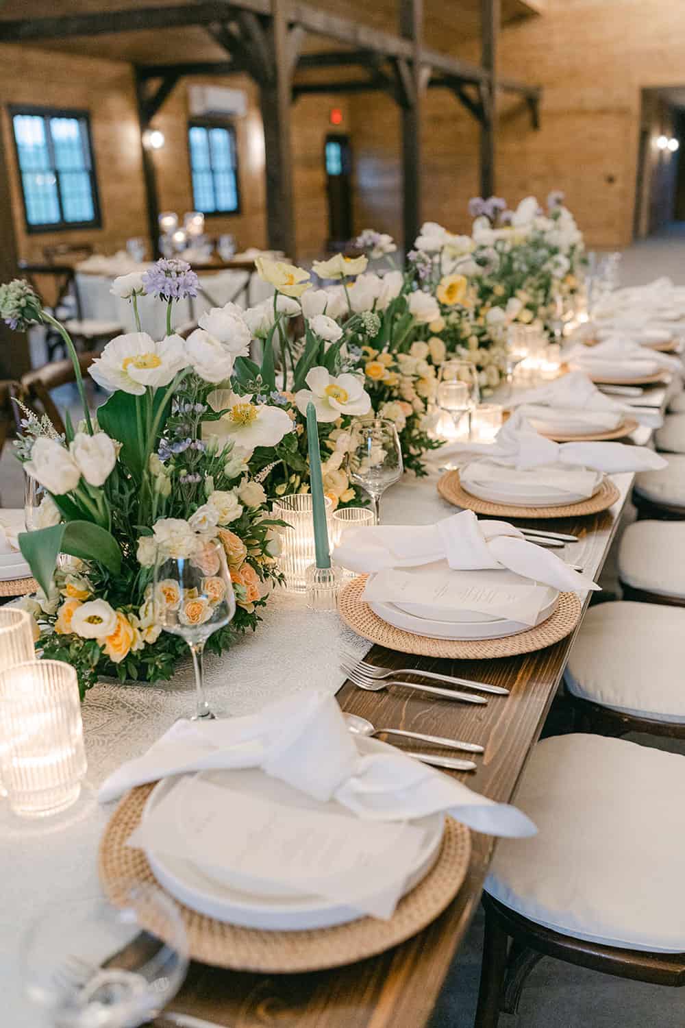 Elegant wedding reception table, set with white and pastel floral centerpieces, candles, and place settings in a rustic venue.