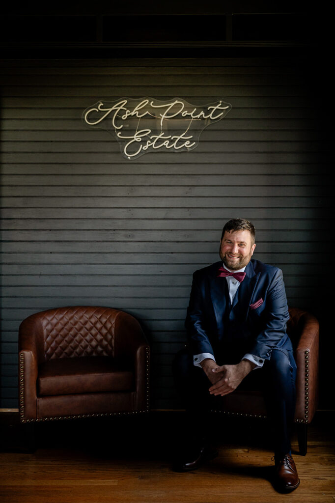 Ash point estate boothbay maine, groom sits in brown leather chair with ash point estate sign lit up above him