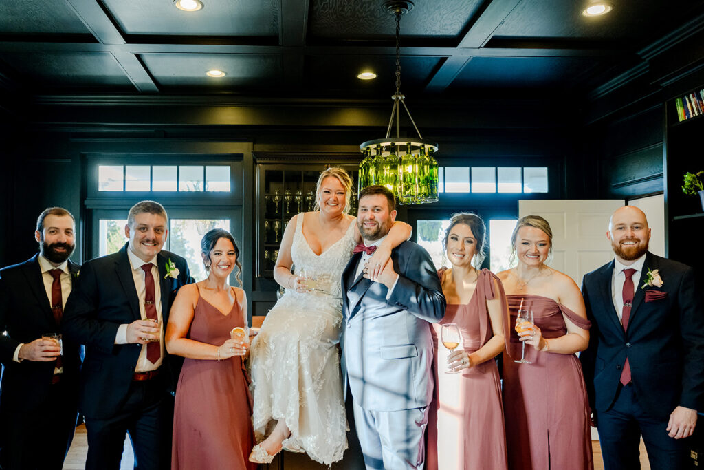 Bride and groom sit on bar top at ash point estate surrounded with bridal party