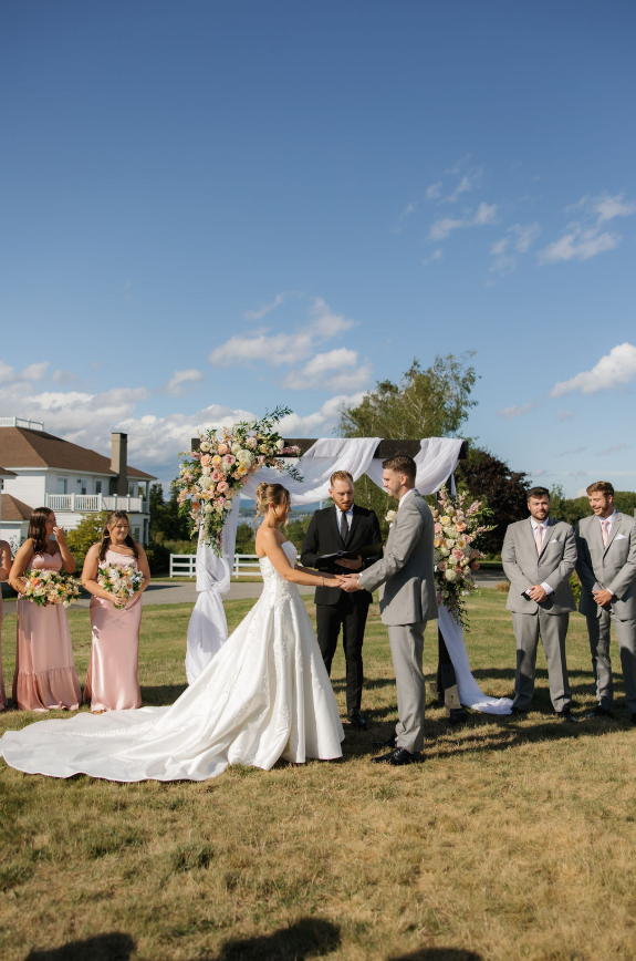 Ash point estate reception venues in maine, newlyweds exchange vows during ceremony at ash point estate, a floral arch behind them